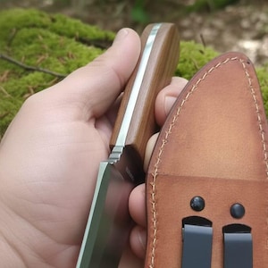 May include: A handcrafted knife with a wooden handle and a stainless steel blade. The knife is held next to a brown leather sheath with two black belt clips. The background is a natural outdoor setting.