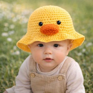 Puede incluir: Un gorro de bebé amarillo tejido a ganchillo con forma de pato, con ojos negros y pico naranja. El gorro lo lleva un bebé con un mono beige, sobre un fondo verde borroso.
