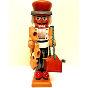 May include: A wooden nutcracker figure wearing a tall orange hat, a white jacket with black buttons, and black trousers. The figure holds a small wooden toolbox with green rope handles and has several miniature wooden clogs hanging from its arm.