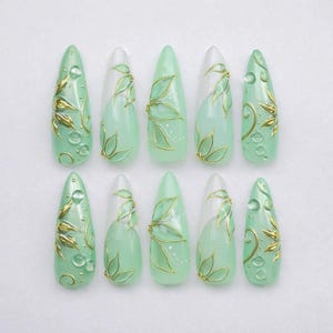 May include: Set of ten almond-shaped false nails. Each nail is painted in a light green hue with gold leaf and clear droplet accents. Some nails feature a white section. The nails are arranged on a white surface.