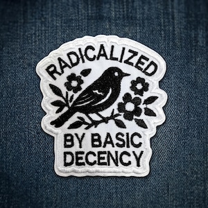 May include: White embroidered patch with a black bird perched on a floral branch. The patch reads "RADICALIZED" above and "BY BASIC DECENCY" below in black capital letters. The patch is on a blue denim background.
