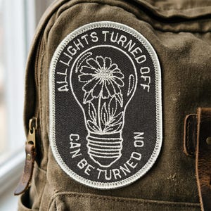 May include: An oval-shaped embroidered patch featuring a black background with white stitching. The design depicts a lightbulb containing a flower, surrounded by the text ALL LIGHTS TURNED OFF CAN BE TURNED ON in a circular white border.