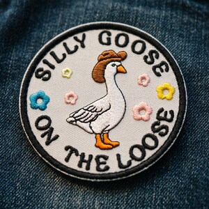 May include: Embroidered patch featuring a white goose wearing a brown cowboy hat and orange boots. The patch is round with the words "SILLY GOOSE ON THE LOOSE" in black. Colorful embroidered flowers surround the goose on a light gray background.