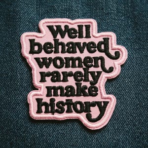 May include: A pink embroidered patch with the text "Well behaved women rarely make history" in black script. The patch has a scalloped edge and is set against a blue denim background.