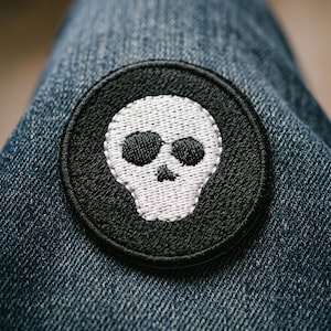 May include: A round, black patch with a white skull design. The skull has large eye sockets and a small nose. The patch is sewn onto blue denim fabric. The patch is a decorative item.