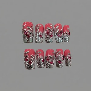 May include: A set of ten artificial nails. Each nail is a vibrant coral color, adorned with silver-colored, ornate designs and small, red gemstones. The nails are arranged in two rows against a neutral gray background, showcasing a glamorous aesthetic.