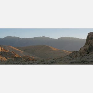 May include: A panoramic landscape view of a desert valley featuring golden-brown rolling hills and rocky outcrops in the foreground, set against a backdrop of distant, hazy blue mountains under a clear, pale sky during the golden hour.