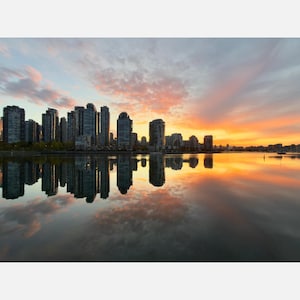 May include: A city skyline featuring numerous high-rise buildings silhouetted against a vibrant sunset with orange, pink, and blue clouds. The buildings and the colourful sky are reflected clearly in the calm, still water in the foreground.