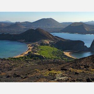 Dark rocky hills around two blue bays, with sandy beaches, green scrub and a tall pointed rock on the right.