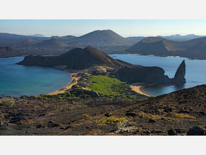 Dark rocky hills around two blue bays, with sandy beaches, green scrub and a tall pointed rock on the right.