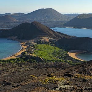 Complete landscape photograph of dark volcanic ridges, two blue bays, sandy beaches and a pointed rock formation on the right.