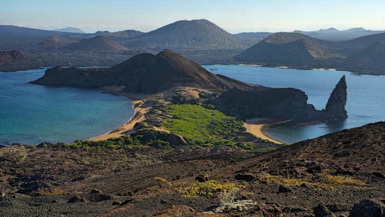 Complete landscape photograph of dark volcanic ridges, two blue bays, sandy beaches and a pointed rock formation on the right.