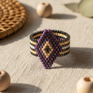 May include: A beaded ring with a geometric design. The ring features a central diamond shape in purple and cream, bordered by black, cream, and white stripes. The ring is set against a neutral background with wooden beads.
