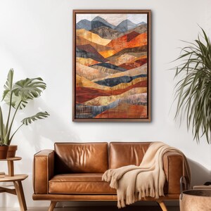 May include: Abstract landscape art print in a wooden frame. The artwork features layered, textured shapes in shades of orange, brown, and blue, depicting a mountain range. The framed art hangs above a brown leather sofa with a beige throw blanket.