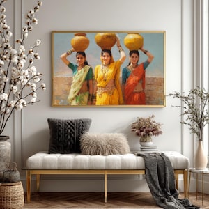 May include: A framed artwork depicts three women in colorful saris carrying pots on their heads. The women are set against a backdrop of a light blue sky and a tan wall. The artwork is displayed above a bench with gold legs, a gray throw, and decorative pillows.