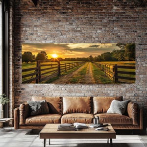 May include: A landscape canvas print depicting a sunset over a dirt road bordered by wooden fences. The artwork hangs above a brown leather sofa with gray pillows, a wooden coffee table, and a brick wall.