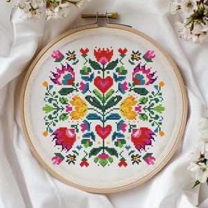 May include: A colorful cross-stitch embroidery in a wooden hoop. The design features a symmetrical floral pattern with red hearts, yellow flowers, and green leaves. The background is white fabric, and the image is framed by white blossoms.