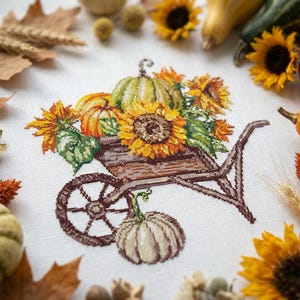 May include: A cross-stitch design depicting a wooden wheelbarrow laden with sunflowers, pumpkins, and gourds. The design is framed by autumn leaves, sunflowers, and small pumpkins. The colour scheme features browns, oranges, yellows, and greens.