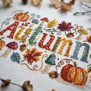 K&ouml;nnte beinhalten: Eine Kreuzstichstickerei auf wei&szlig;em Stoff mit dem Wort AUTUMN in gro&szlig;en, bunten Blockbuchstaben. Das Design ist mit orangefarbenen K&uuml;rbissen, verschiedenen Pilzen, roten und gelben Ahornbl&auml;ttern sowie kleinen braunen Eicheln verziert.