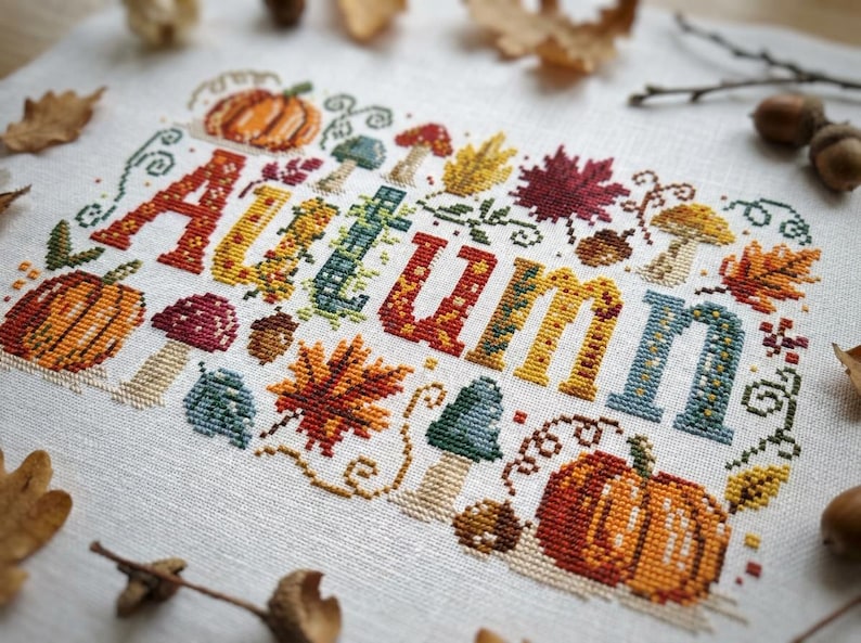 K&ouml;nnte beinhalten: Eine Kreuzstichstickerei auf wei&szlig;em Stoff mit dem Wort AUTUMN in gro&szlig;en, bunten Blockbuchstaben. Das Design ist mit orangefarbenen K&uuml;rbissen, verschiedenen Pilzen, roten und gelben Ahornbl&auml;ttern sowie kleinen braunen Eicheln verziert.