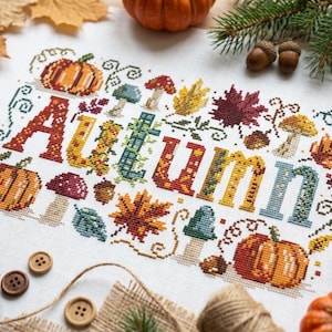 K&ouml;nnte beinhalten: Ein wei&szlig;es Stoffst&uuml;ck mit einem farbenfrohen Kreuzstichmuster, das das Wort Autumn in gro&szlig;en, gemusterten Buchstaben zeigt. Die Stickerei umfasst orangefarbene K&uuml;rbisse, rote und gelbe Ahornbl&auml;tter, braune Eicheln sowie verschiedene blaue und rote Pilze.