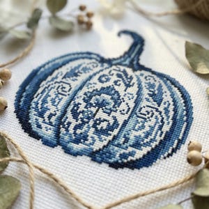 May include: A cross-stitch design of a blue and white pumpkin. The pumpkin features a detailed floral pattern. The stem is a darker blue. The design is on white fabric, with dried leaves and twine in the background, creating a rustic aesthetic.