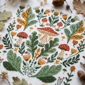 Könnte beinhalten: Eine Kreuzstichstickerei auf weißem Stoff mit einem botanischen Muster aus rotköpfigen Pilzen, grünen Eichenblättern und kleinen gelben Blumen. Das Design ist in einer dichten, symmetrischen Anordnung mit verschiedenen Waldmotiven gestaltet.