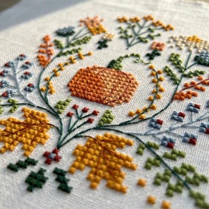 K&ouml;nnte beinhalten: Eine Kreuzstichstickerei auf wei&szlig;em Stoff mit einem zentralen orangefarbenen K&uuml;rbis, umgeben von einer kreisf&ouml;rmigen Anordnung herbstlicher botanischer Elemente, darunter gelbe Ahornbl&auml;tter, gr&uuml;ne Zweige sowie kleine blaue und orangefarbene Bl&uuml;ten.