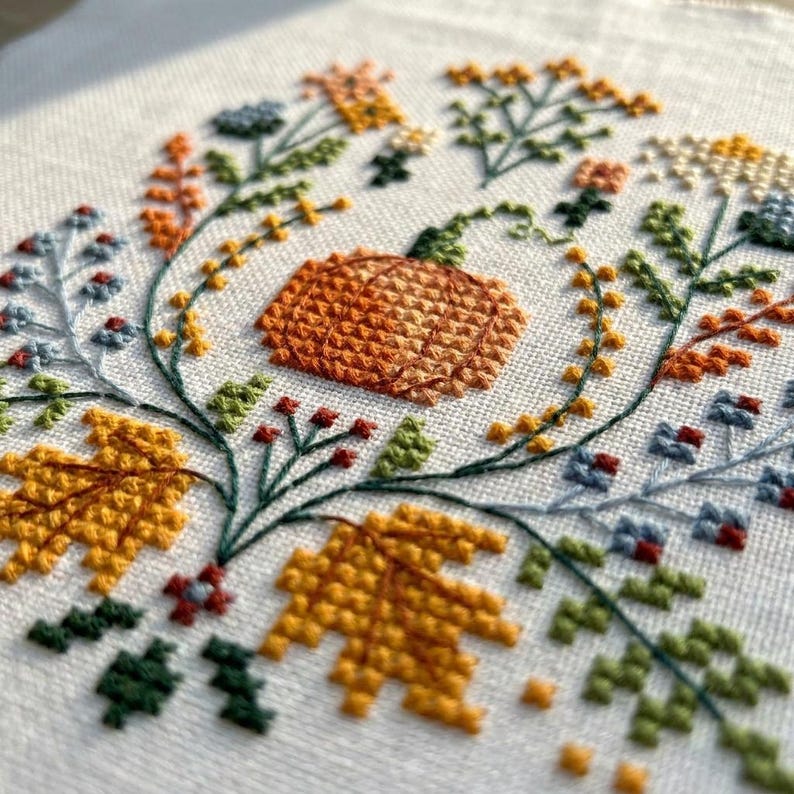 K&ouml;nnte beinhalten: Eine Kreuzstichstickerei auf wei&szlig;em Stoff mit einem zentralen orangefarbenen K&uuml;rbis, umgeben von einer kreisf&ouml;rmigen Anordnung herbstlicher botanischer Elemente, darunter gelbe Ahornbl&auml;tter, gr&uuml;ne Zweige sowie kleine blaue und orangefarbene Bl&uuml;ten.