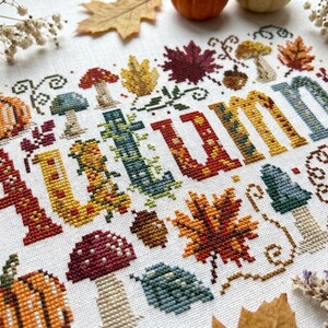 K&ouml;nnte beinhalten: Eine Kreuzstichstickerei auf wei&szlig;em Stoff mit dem Wort AUTUMN in gro&szlig;en, bunten Blockbuchstaben. Das Design ist umgeben von gestickten Motiven aus orangefarbenen K&uuml;rbissen, roten und gelben Ahornbl&auml;ttern, braunen Eicheln und verschiedenen Pilzen.