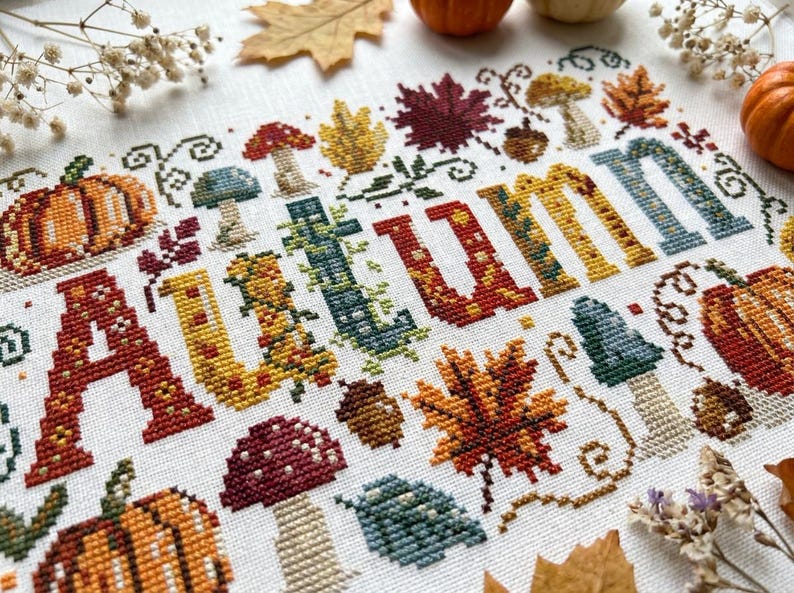 K&ouml;nnte beinhalten: Eine Kreuzstichstickerei auf wei&szlig;em Stoff mit dem Wort AUTUMN in gro&szlig;en, bunten Blockbuchstaben. Das Design ist umgeben von gestickten Motiven aus orangefarbenen K&uuml;rbissen, roten und gelben Ahornbl&auml;ttern, braunen Eicheln und verschiedenen Pilzen.