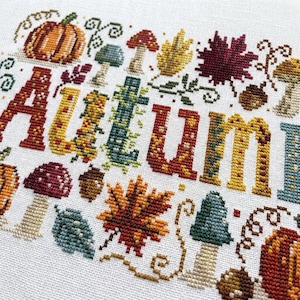 K&ouml;nnte beinhalten: Eine Kreuzstichstickerei auf wei&szlig;em Stoff mit dem Wort AUTUMN in gro&szlig;en, dekorativen, mehrfarbigen Buchstaben. Das Design ist umgeben von gestickten Motiven aus orangefarbenen K&uuml;rbissen, roten und gelben Ahornbl&auml;ttern, braunen Eicheln und Pilzen.