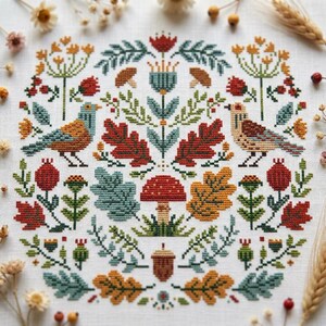 May include: A cross-stitch embroidery pattern on white fabric featuring two birds facing each other, surrounded by a symmetrical arrangement of red and yellow oak leaves, green foliage, red mushrooms, berries, and a central brown acorn.