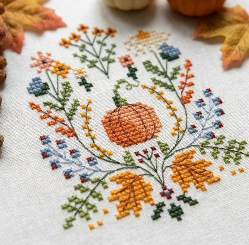 K&ouml;nnte beinhalten: Eine Kreuzstichstickerei auf wei&szlig;em Stoff mit einem zentralen orangefarbenen K&uuml;rbis, umgeben von einem farbenfrohen Arrangement herbstlicher botanischer Elemente, darunter gelbe und orangefarbene Ahornbl&auml;tter, kleine blaue Blumen und gr&uuml;nes Blattwerk.