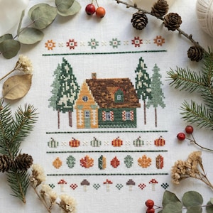 May include: Cross-stitch embroidery depicting a house with a brown roof and green and yellow walls, with green trees on either side. The design includes pumpkins, leaves, and mushrooms, with a border of floral and geometric patterns. The artwork is set against a white background.
