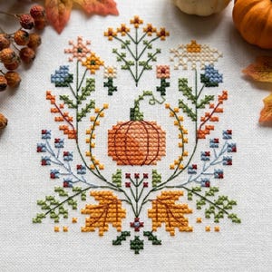 K&ouml;nnte beinhalten: Eine Kreuzstichstickerei auf wei&szlig;em Stoff mit einem zentralen orangefarbenen K&uuml;rbis, umgeben von einer symmetrischen Anordnung aus bunten Blumen, gr&uuml;nen Stielen und goldgelben Herbstbl&auml;ttern in Blau-, Orange- und Gelbt&ouml;nen.