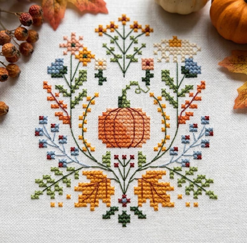 K&ouml;nnte beinhalten: Eine Kreuzstichstickerei auf wei&szlig;em Stoff mit einem zentralen orangefarbenen K&uuml;rbis, umgeben von einer symmetrischen Anordnung aus bunten Blumen, gr&uuml;nen Stielen und goldgelben Herbstbl&auml;ttern in Blau-, Orange- und Gelbt&ouml;nen.