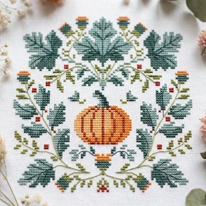 Peut inclure: Une broderie au point de croix sur tissu blanc représentant une citrouille orange centrale entourée d'un arrangement symétrique de feuilles vert foncé, de petites fleurs orange et de minuscules accents carrés rouges.