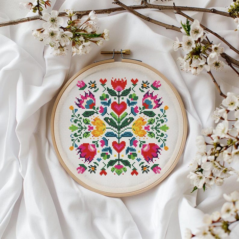 May include: A colorful cross-stitch embroidery in a wooden hoop. The design features a symmetrical floral pattern with red hearts, yellow flowers, and green leaves. The background is white fabric, and the image is framed by white blossoms.