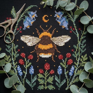 May include: Cross-stitch embroidery on black fabric featuring a large bumblebee with detailed wings, surrounded by a floral wreath. The design includes red berries, blue flowers, and a crescent moon. A pair of silver scissors is placed near the top.