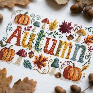 May include: A cross-stitch embroidery on white fabric featuring the word Autumn in large, multicoloured block letters. The design is surrounded by various seasonal motifs including orange pumpkins, red and yellow mushrooms, and brown maple leaves.