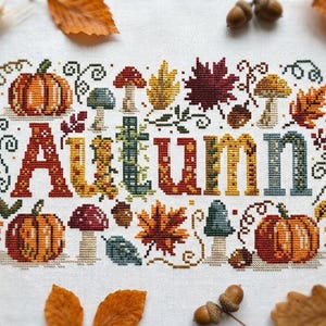 K&ouml;nnte beinhalten: Eine Kreuzstichstickerei auf wei&szlig;em Stoff mit dem Wort Autumn in gro&szlig;en, bunten Buchstaben. Das Design ist von verschiedenen herbstlichen Motiven umgeben, darunter orangefarbene K&uuml;rbisse, rote und braune Pilze, Eicheln und bunte Ahornbl&auml;tter.