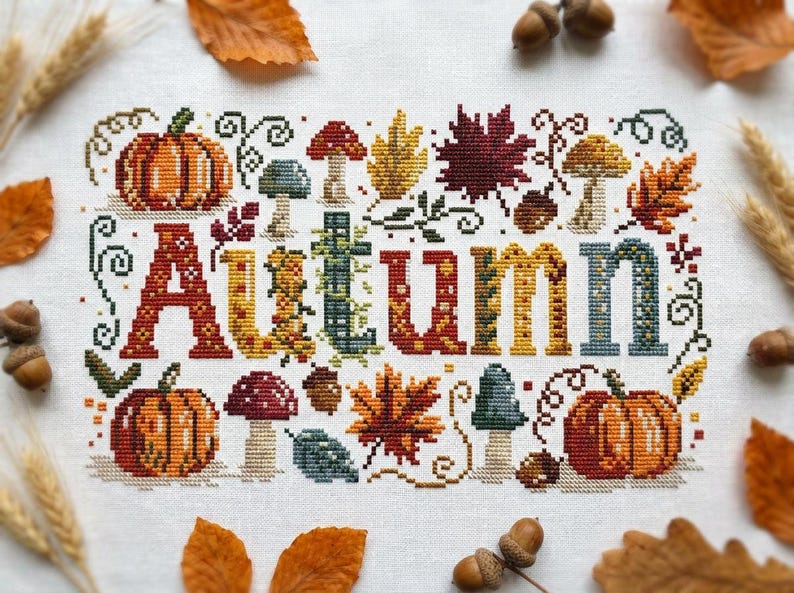K&ouml;nnte beinhalten: Eine Kreuzstichstickerei auf wei&szlig;em Stoff mit dem Wort Autumn in gro&szlig;en, bunten Buchstaben. Das Design ist von verschiedenen herbstlichen Motiven umgeben, darunter orangefarbene K&uuml;rbisse, rote und braune Pilze, Eicheln und bunte Ahornbl&auml;tter.
