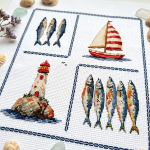 May include: A white fabric piece featuring four distinct cross-stitch sections bordered by dark blue lines. The designs include three blue fish, a red and white sailboat, a lighthouse on a rocky base, and five colourful fish in a row.