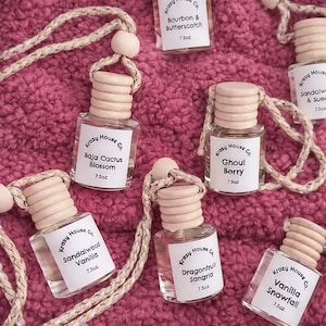 May include: Several small glass bottles with wooden lids and rope cords, each labelled with a scent name like "Dragonfruit Sangria". The bottles, containing 7.5oz of product, are arranged on a pink, textured surface. The labels feature the brand "Krazy House Co."
