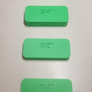 May include: Four green rectangular blocks with measurements: 1", 3/4", 1/2", and 1/4". The text "CALENAUS3D" is printed on each block. The blocks are arranged vertically on a light surface.