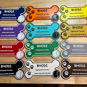 May include: A collection of colorful dog bone-shaped pet tags. Each tag features the name "BHODI", paw prints, a tennis ball graphic, and contact information. The tags come in various colors, including blue, orange, yellow, purple, red, green, gray, black, white, and brown.