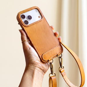 May include: A tan leather phone case with visible white stitching along the edges. The case features a cutout for the camera lenses and is attached to a matching tan leather strap using two antique brass-coloured metal swivel lobster clasps.
