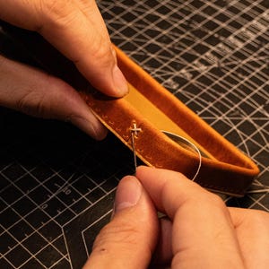 May include: A tan leather tray with stitched edges being assembled with a needle and thread. The leather has a smooth, matte finish and a rectangular shape with rounded corners. A thin, light-colored thread is pulled through the pre-punched holes.