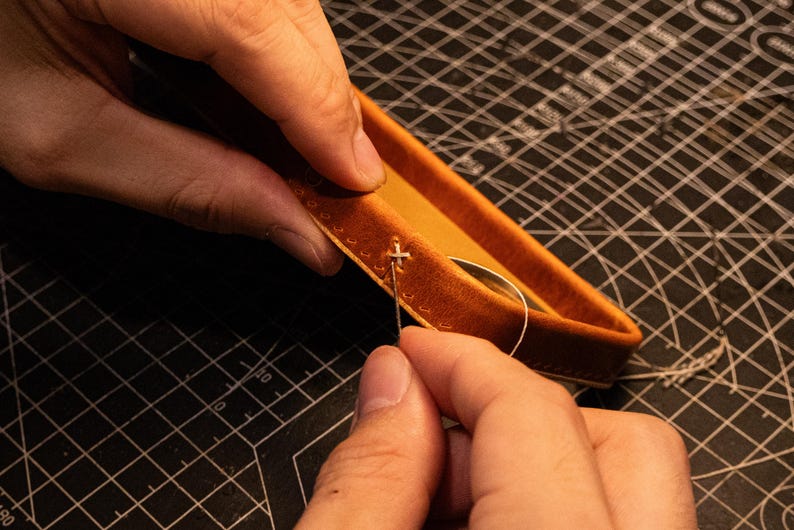 May include: A tan leather tray with stitched edges being assembled with a needle and thread. The leather has a smooth, matte finish and a rectangular shape with rounded corners. A thin, light-colored thread is pulled through the pre-punched holes.